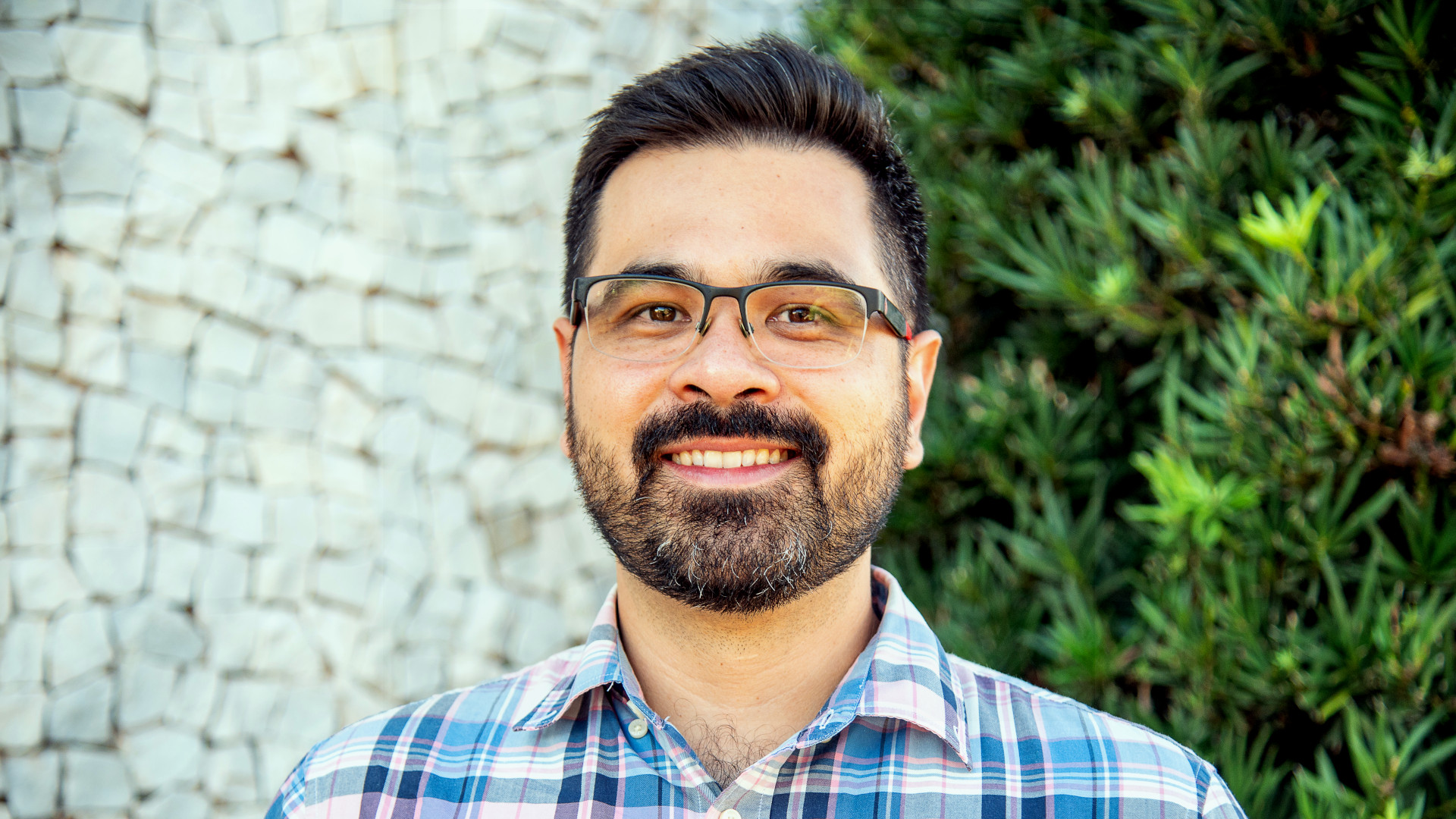 Photo of a man with slight southeast asian features, tan skin, short black hair, a short black beard with some gray hairs, black-rimmed glasses, and a checkered shirt.